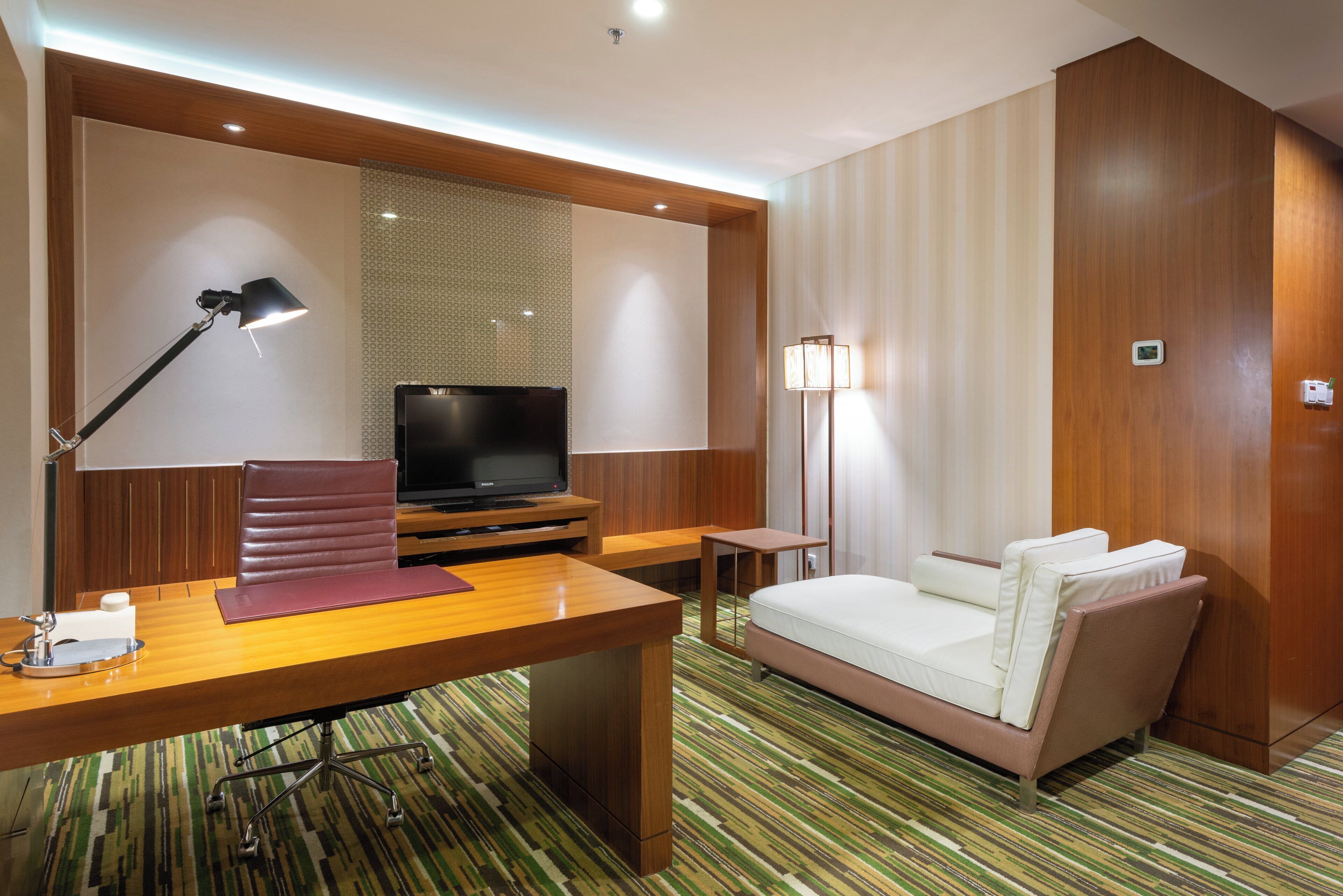 Una habitación de hotel con un escritorio de madera, televisor, sofá, lámpara de pie y alfombra estampada.