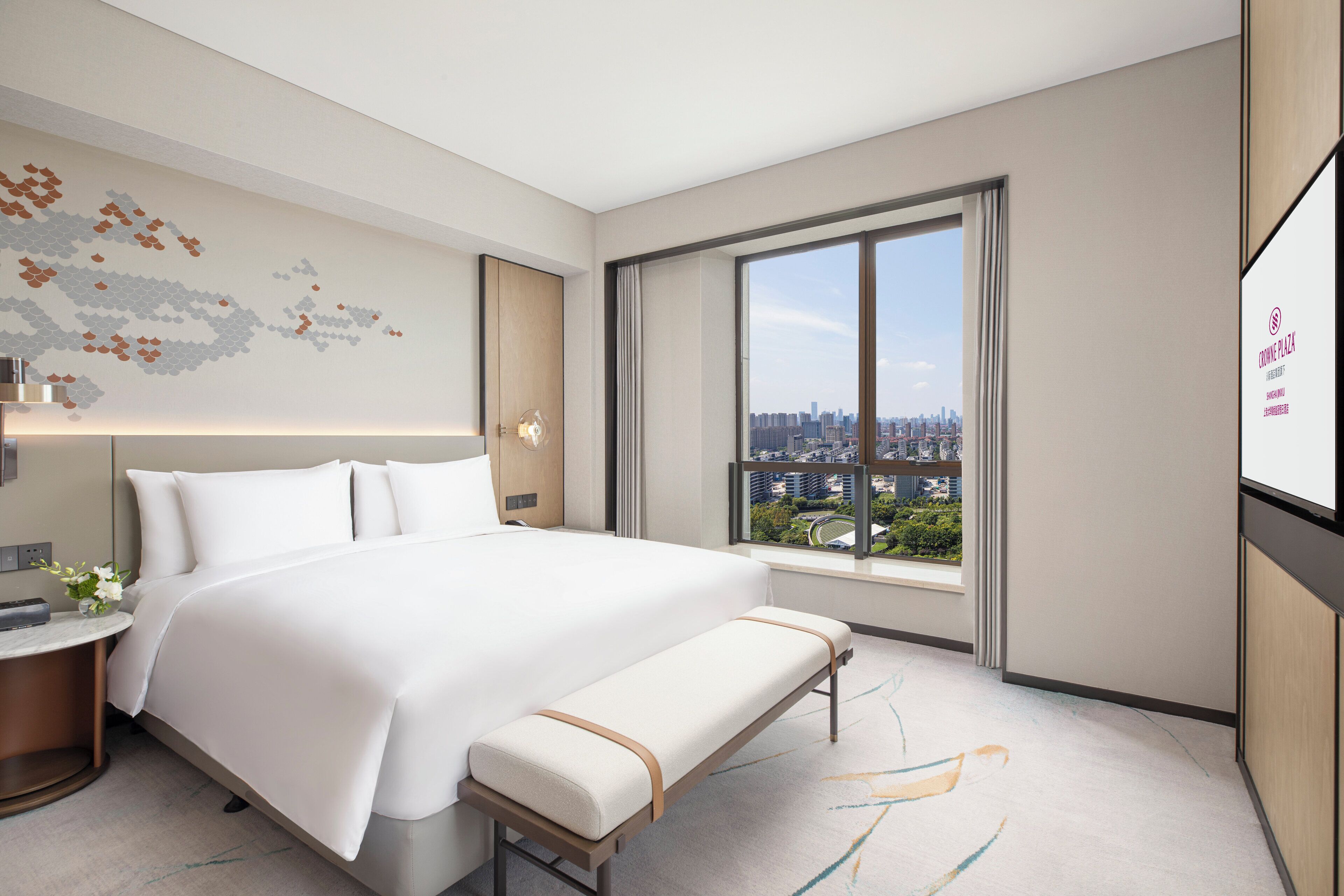 Ein modernes Hotelzimmer mit einem großen Bett, einer Bank, einem runden Tisch mit Blumen und Blick auf die Stadt durch ein großes Fenster.