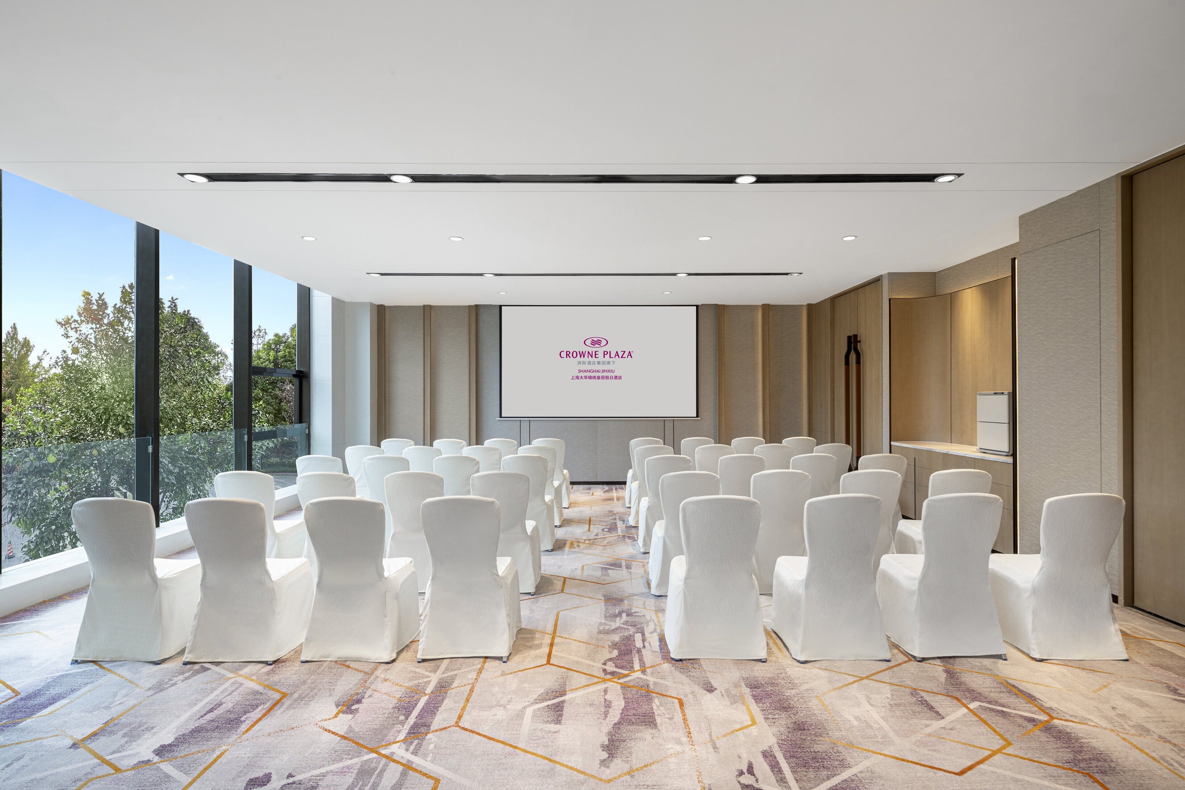 A conference room with rows of white chairs, a large screen displaying text, and a view of trees through large windows.
