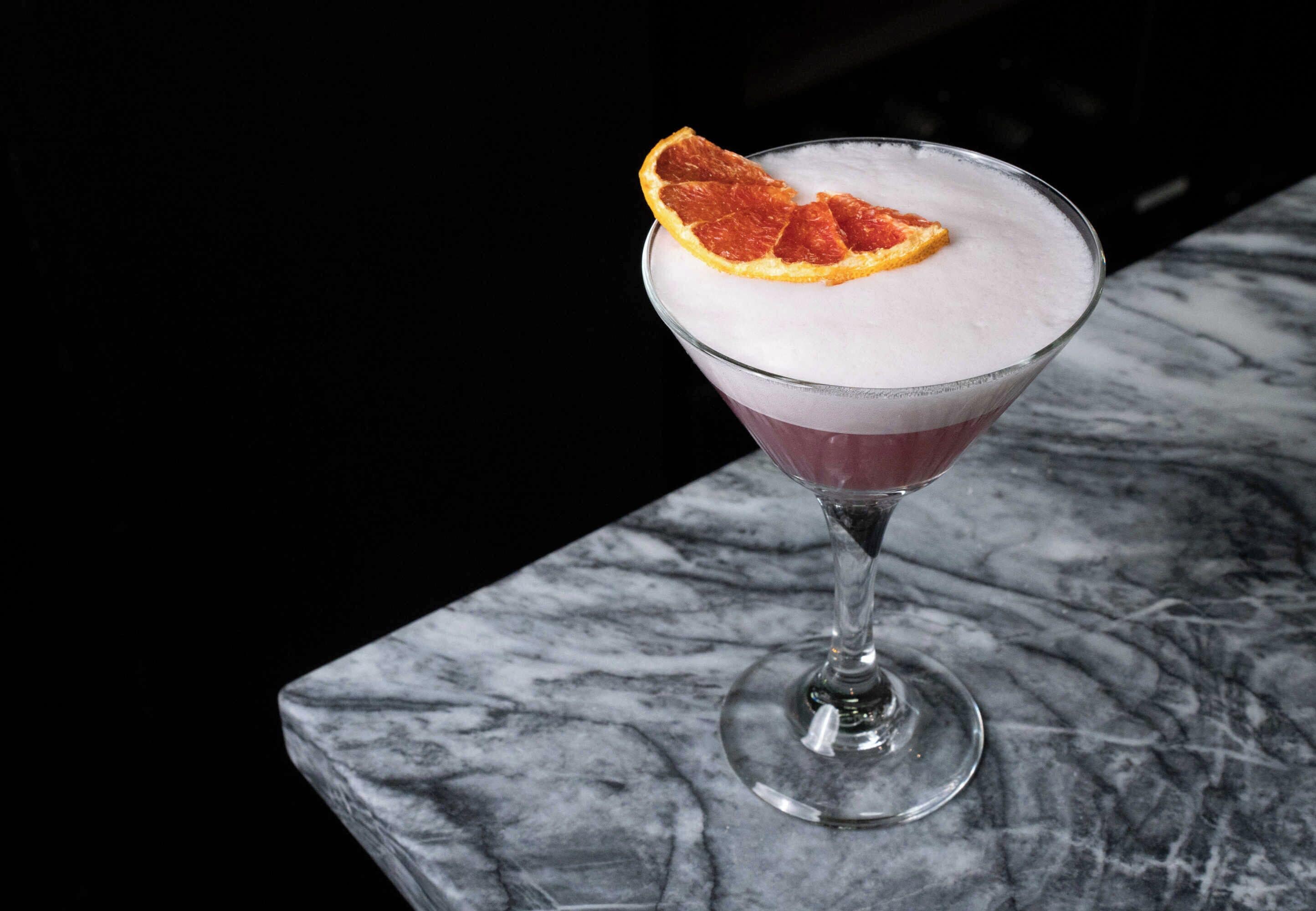 Un verre à martini contenant une boisson mousseuse et décoré d’une tranche d’orange séchée, le tout posé sur une surface en marbre.