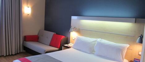 A hotel room with a large bed, a bedside table with a lamp, a chair with red cushions, and a curtain.