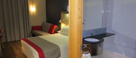 A hotel room with a bed, a bedside table, a lamp, a bathroom with a sink and a mirror.