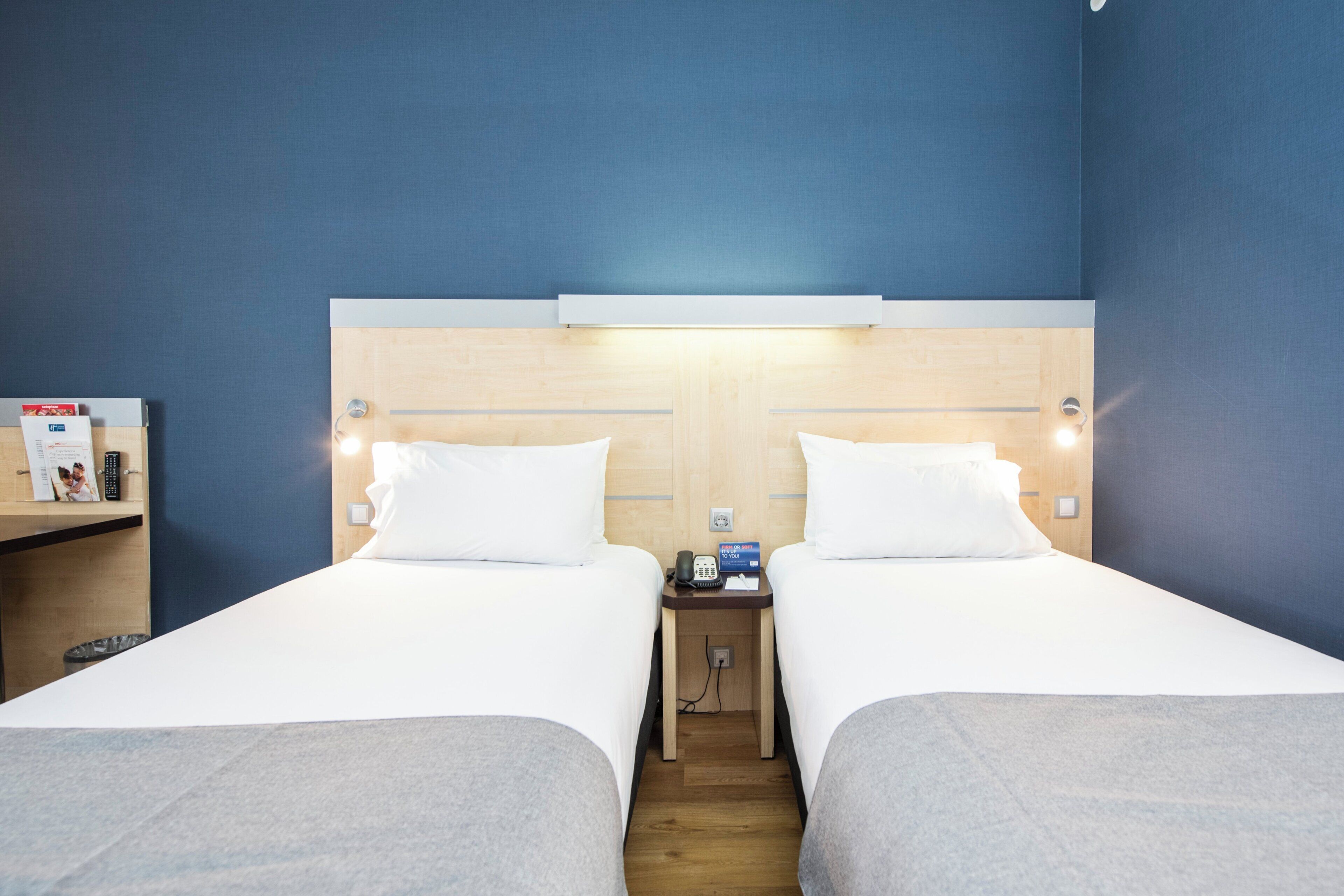 A hotel room with two single beds, a bedside table with a lamp, a small desk with a phone and a sign, and a wall-mounted shelf with items.