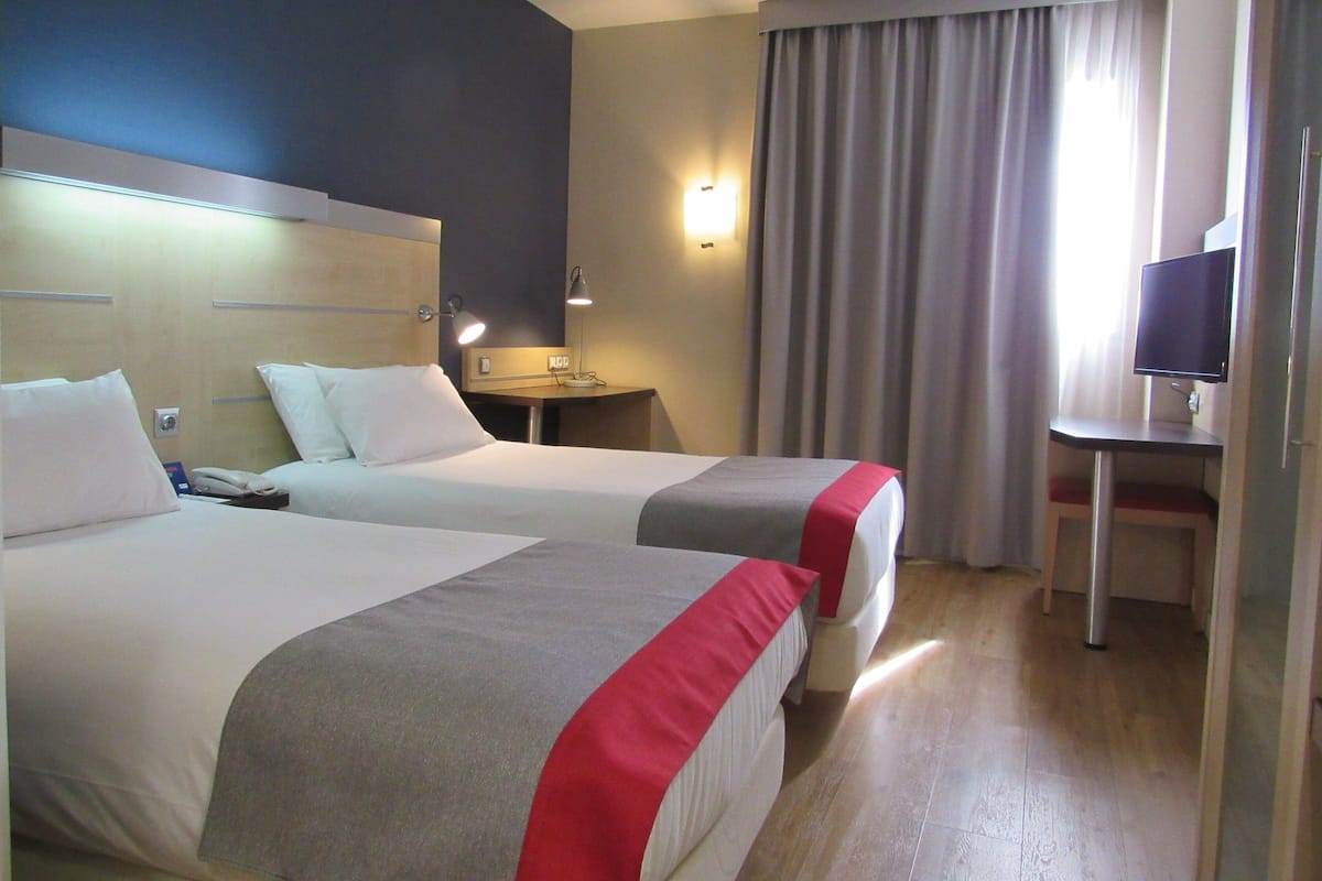 A hotel room with two beds, a desk, a TV, and a window with curtains.