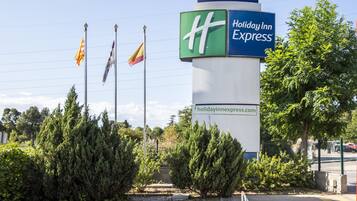 Letrero de Holiday Inn Express con una dirección web, rodeado de árboles de hoja perenne y un cielo despejado.