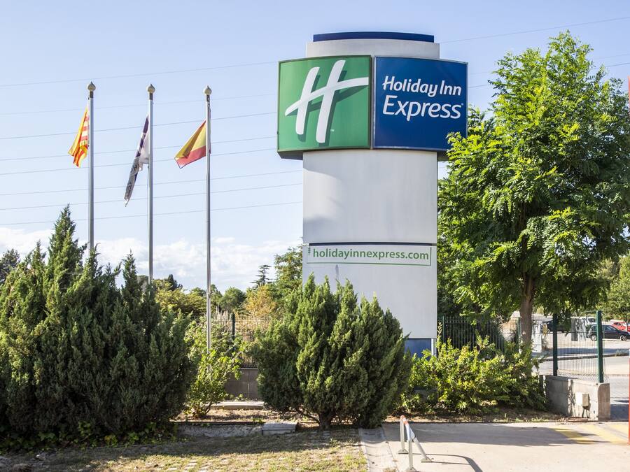 Letrero de Holiday Inn Express con una dirección web, rodeado de árboles de hoja perenne y un cielo despejado.