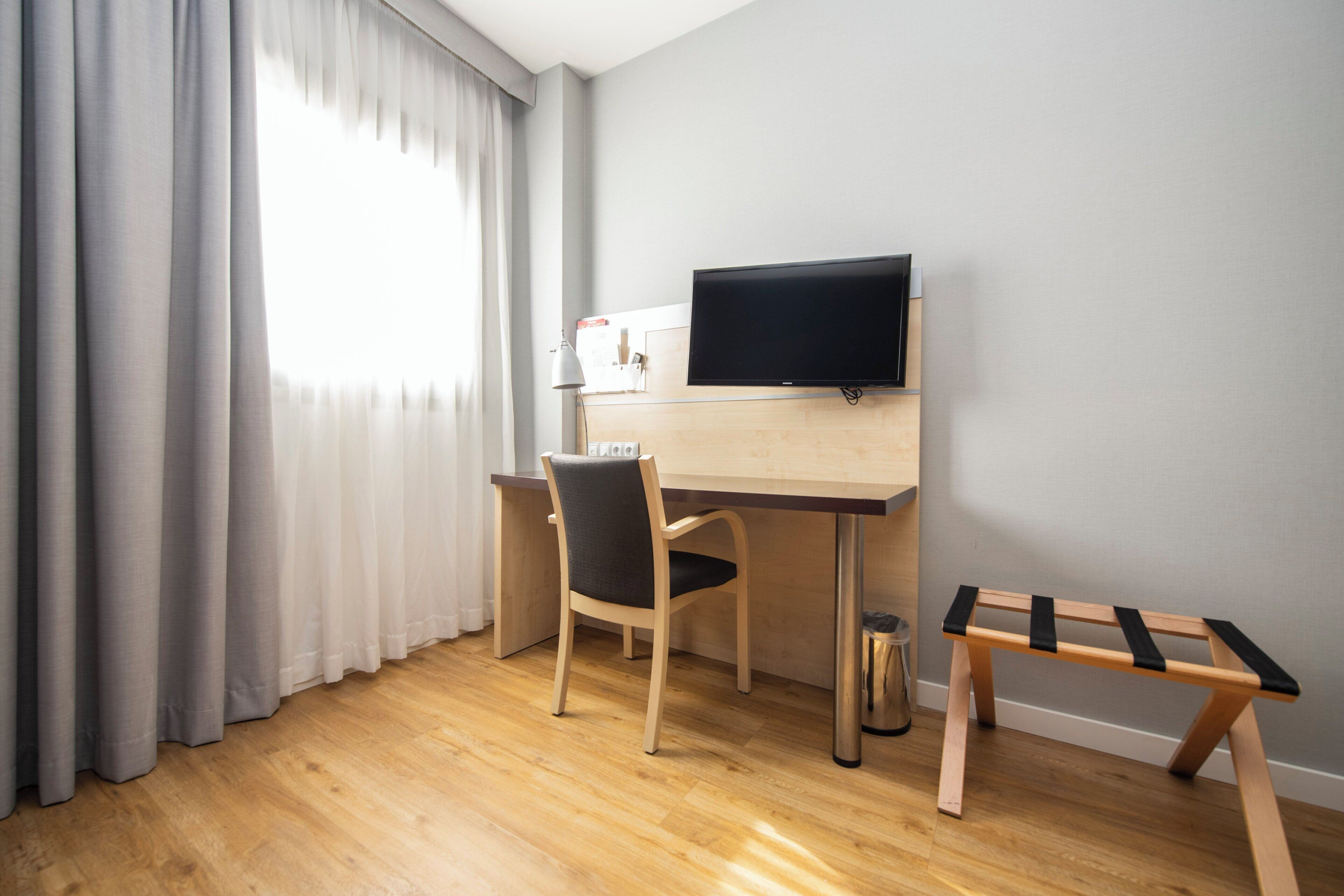 Una habitación con un escritorio de madera, una silla, un televisor montado en la pared, una mesita y una ventana con cortinas.
