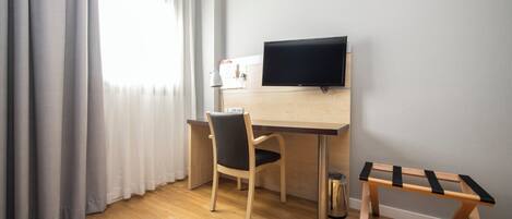 Una habitación con un escritorio de madera, una silla, un televisor montado en la pared, una mesita y una ventana con cortinas.