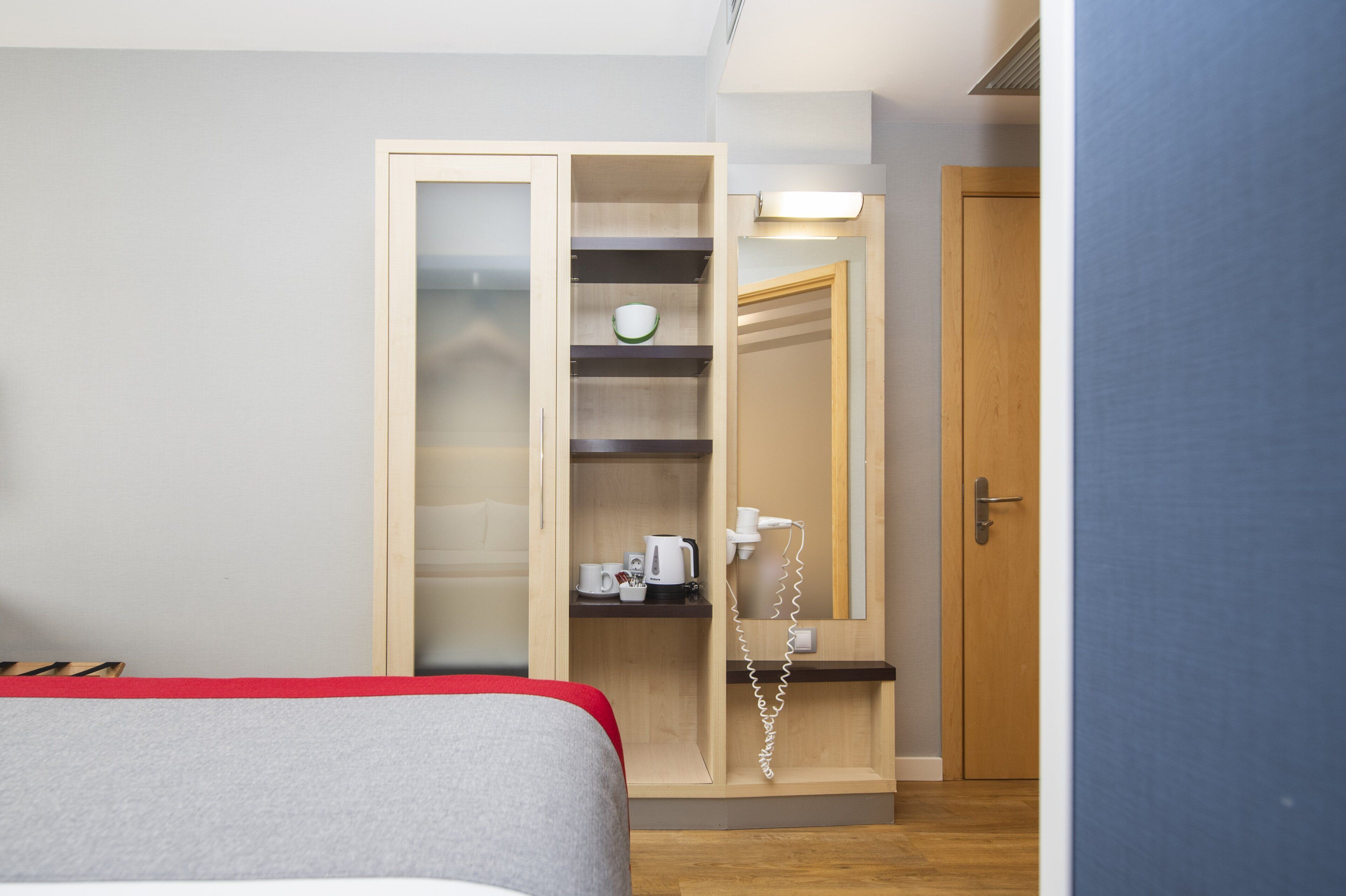 A hotel room with a bed, a wardrobe with a mirror, a telephone, and a shelf with a kettle and cups.