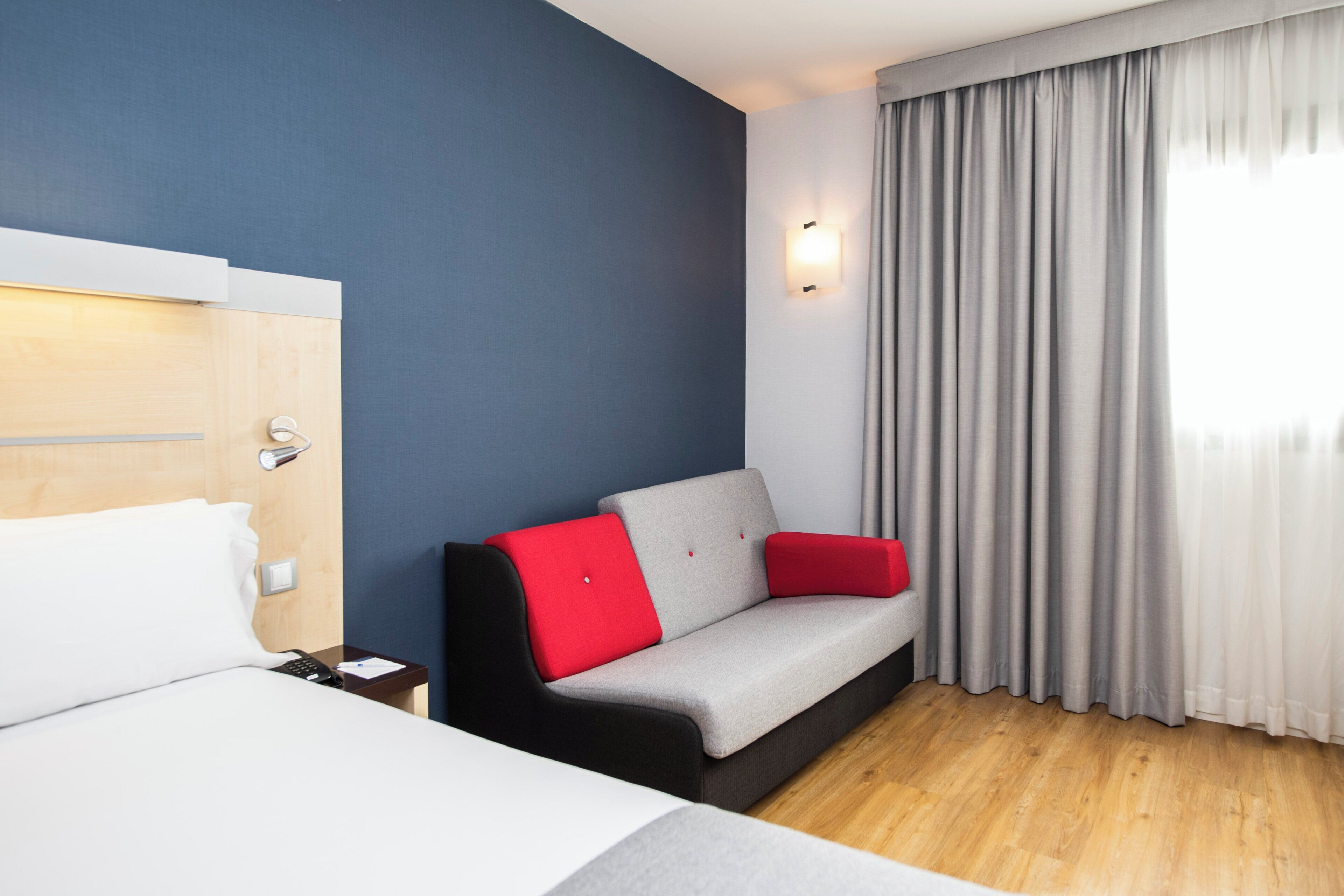 A hotel room with a bed, a sofa with red cushions, a wooden floor, and a window with curtains.