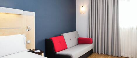 A hotel room with a bed, a sofa with red cushions, a wooden floor, and a window with curtains.