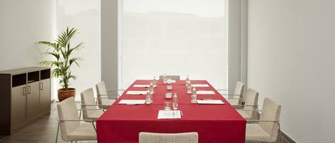Un salón de conferencias con una mesa roja, sillas blancas, una planta en maceta y un ventanal amplio.
