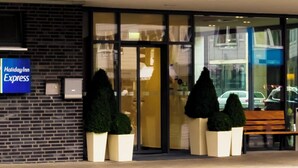 A Holiday Inn Express sign on a brick wall, a glass entrance with a bench outside, and potted plants. - Holiday Inn Express Guetersloh by IHG (Guetersloh)
