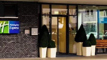 Señal de Holiday Inn Express sobre un muro de ladrillo, una entrada de vidrio con un banco afuera y plantas en macetas.