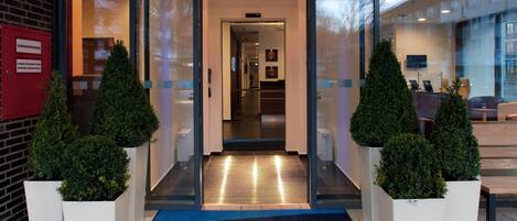 Entrada de un hotel con puerta de vidrio, alfombra azul y dos plantas en macetas.