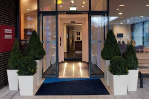 A hotel entrance with a glass door, a blue carpet, and two potted plants. - Holiday Inn Express Guetersloh by IHG (Guetersloh)
