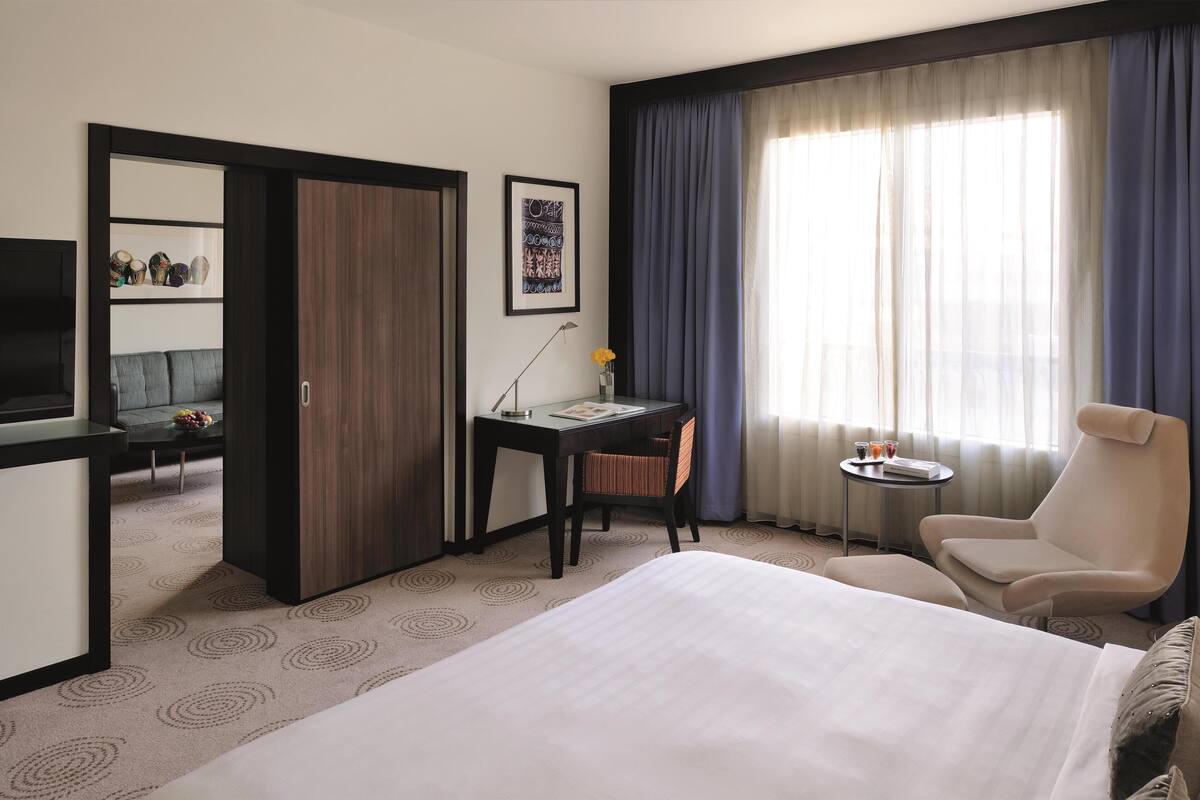 A hotel room with a bed, a desk, a chair, a TV, a small table with a vase of flowers, and a large window with curtains.