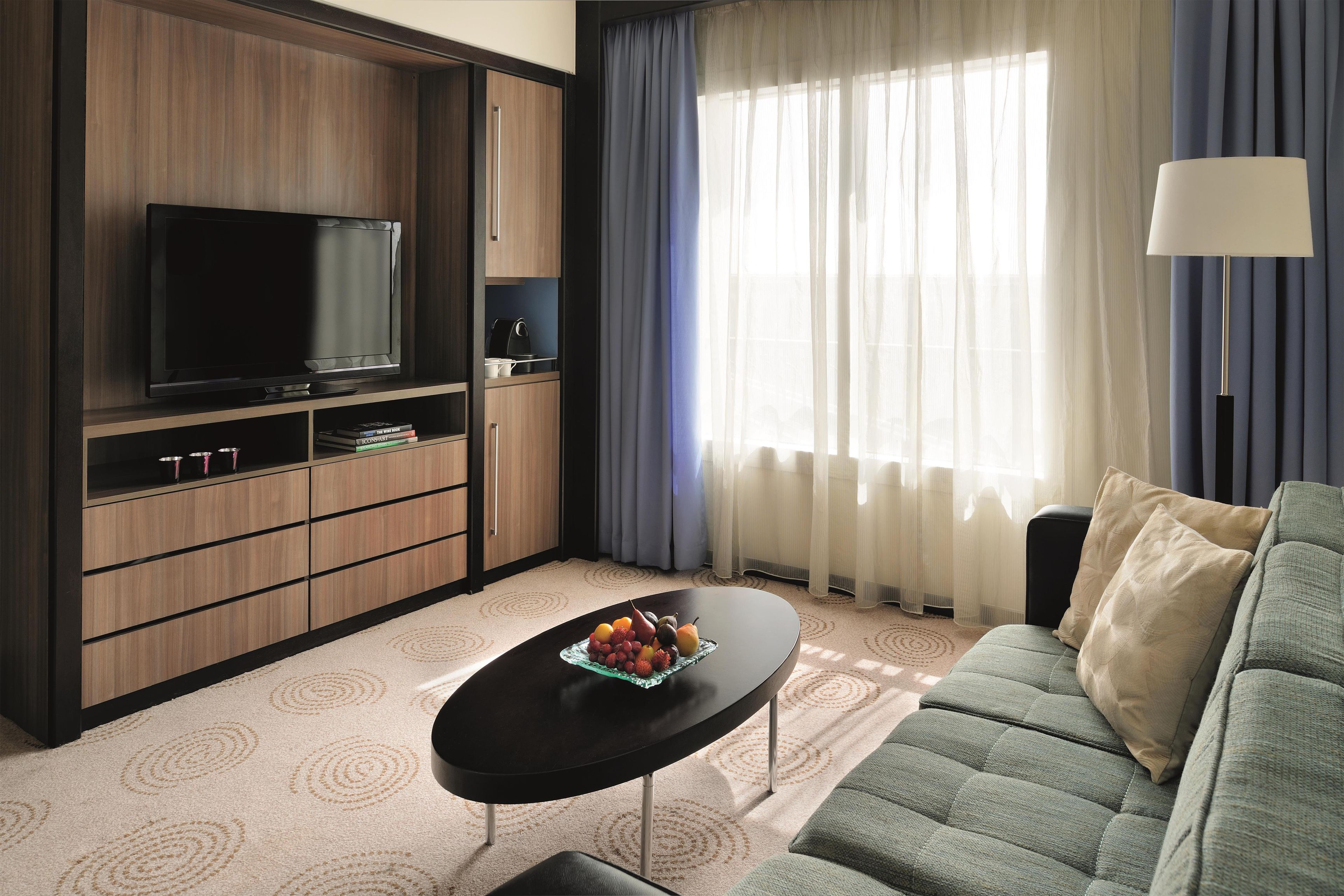 A modern living room with a flat-screen TV, a sofa, a coffee table with fruit, and a large window with curtains.