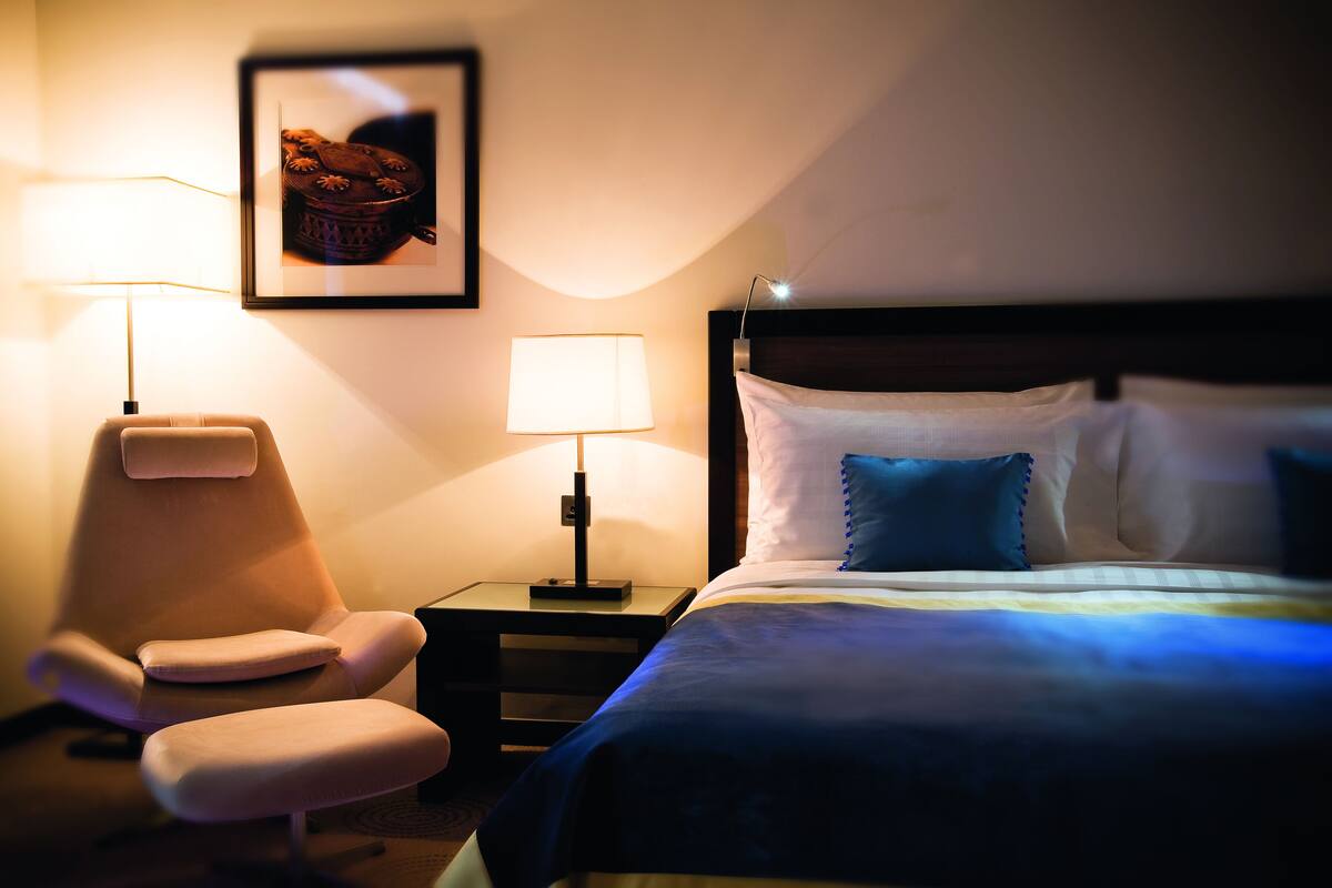 A hotel room with a bed, two bedside lamps, a chair, and a framed picture on the wall.