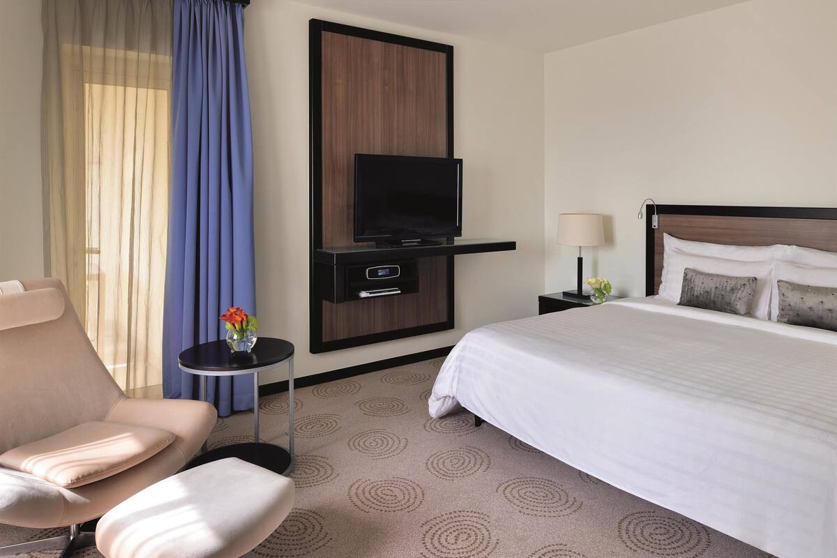 A hotel room with a bed, a TV, a chair, a small table with a vase of flowers, and a nightstand with a lamp.