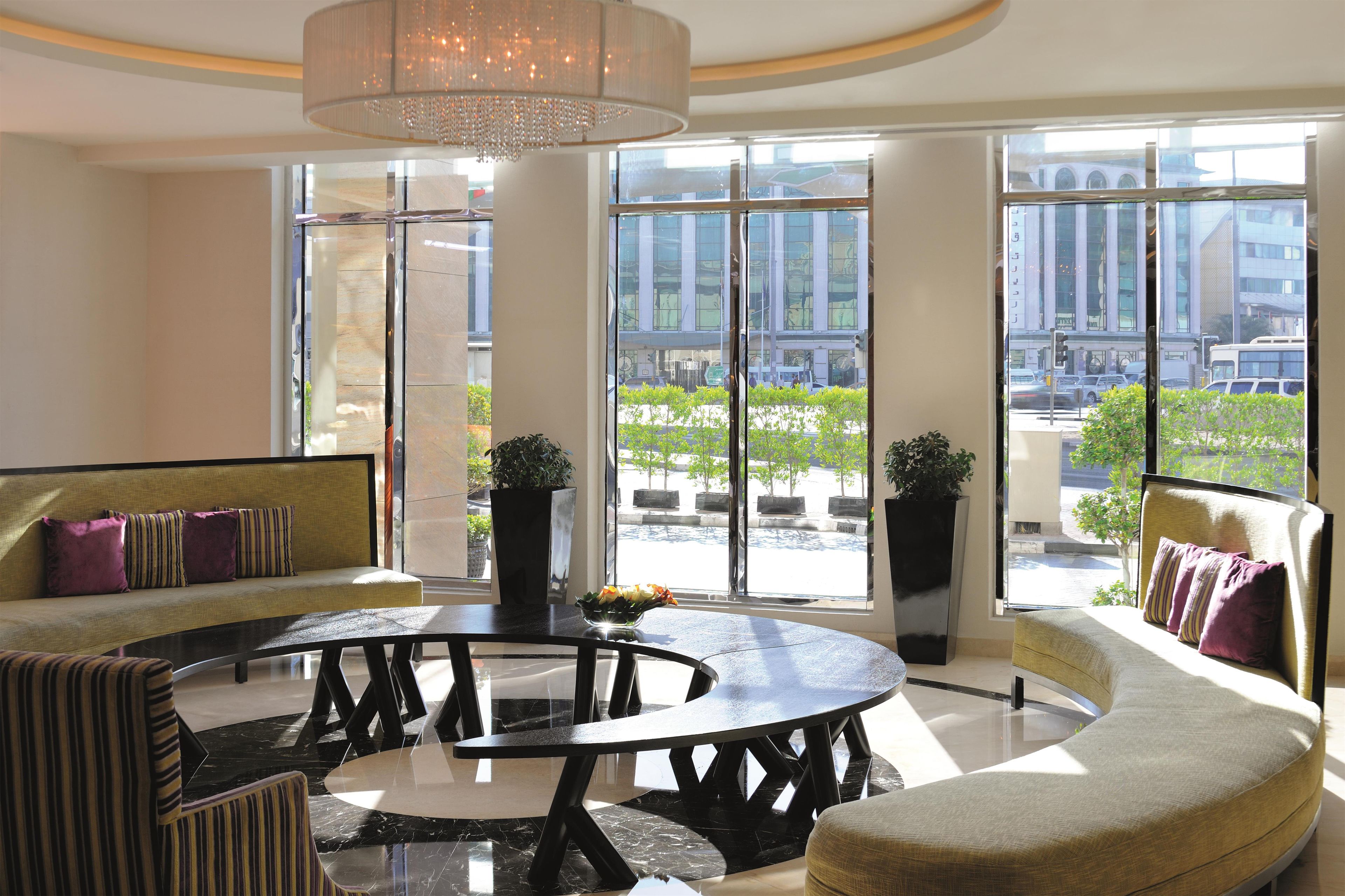 A modern lounge area with a round table, sofas, and large windows offering a view of the cityscape.