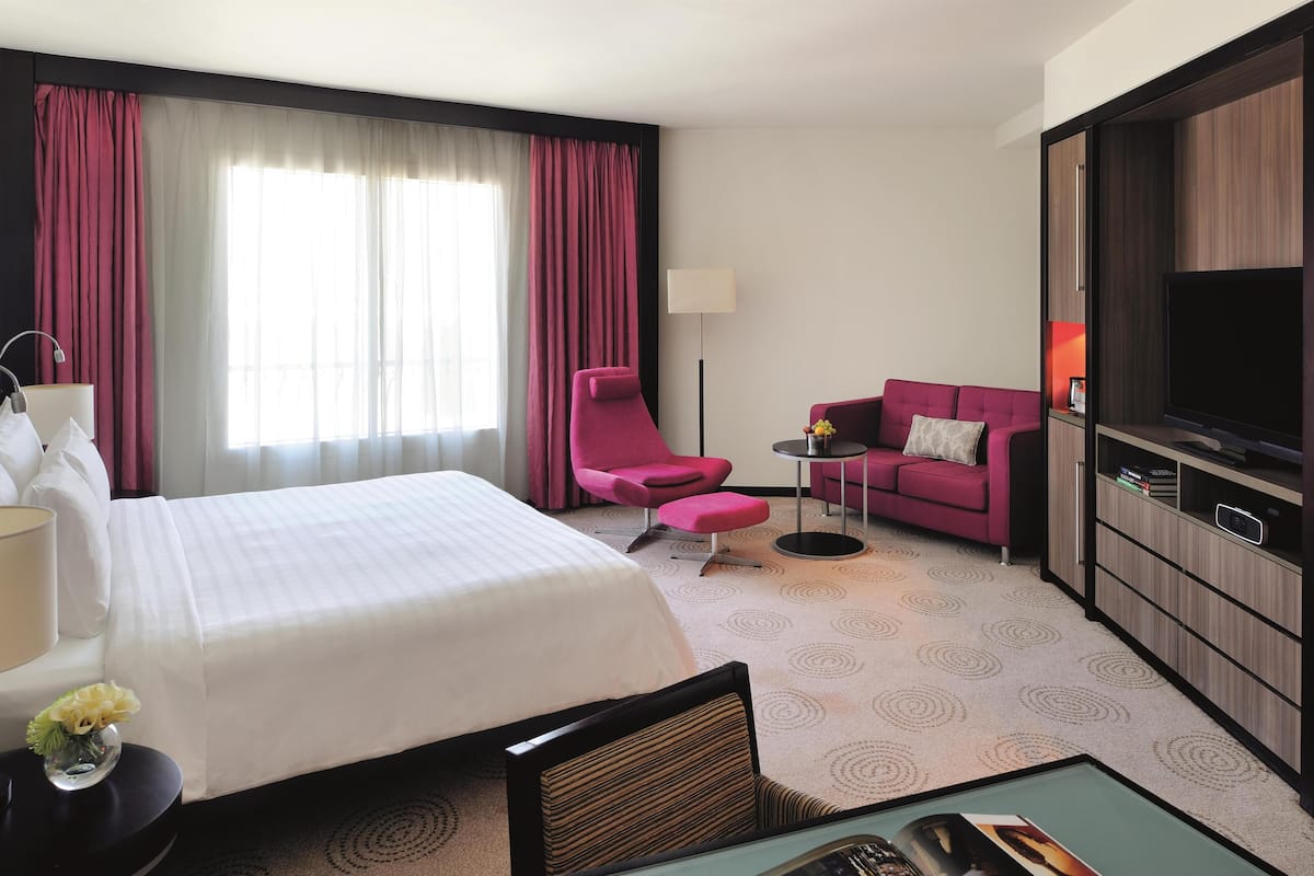 A hotel room with a large bed, a pink armchair, a small sofa, a TV, and a minibar.