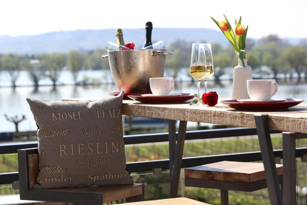 A lakeside dining setup with a table set for two, featuring a wine glass, a bottle of champagne, and tulips in a vase.