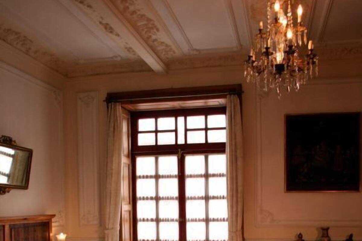 A bedroom with a large bed, a chandelier, a window with curtains, a painting on the wall, and a small table with decorative items.