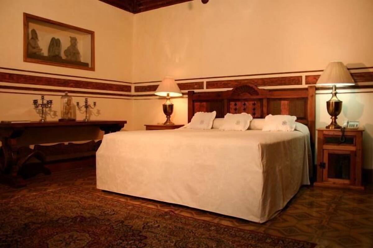 A hotel room with a large bed, two bedside tables with lamps, a wooden headboard, a desk with a chair, and a patterned carpet.
