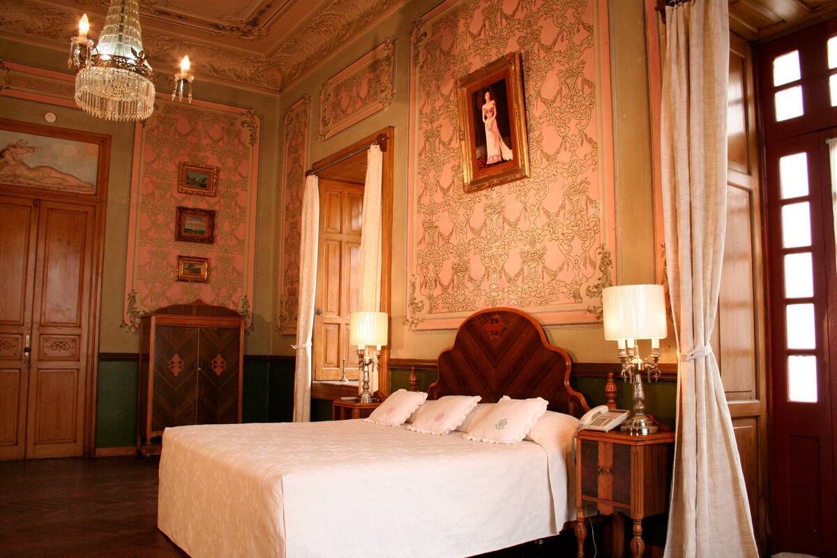 A classic hotel room with a large bed, ornate wallpaper, a chandelier, and a wooden wardrobe.