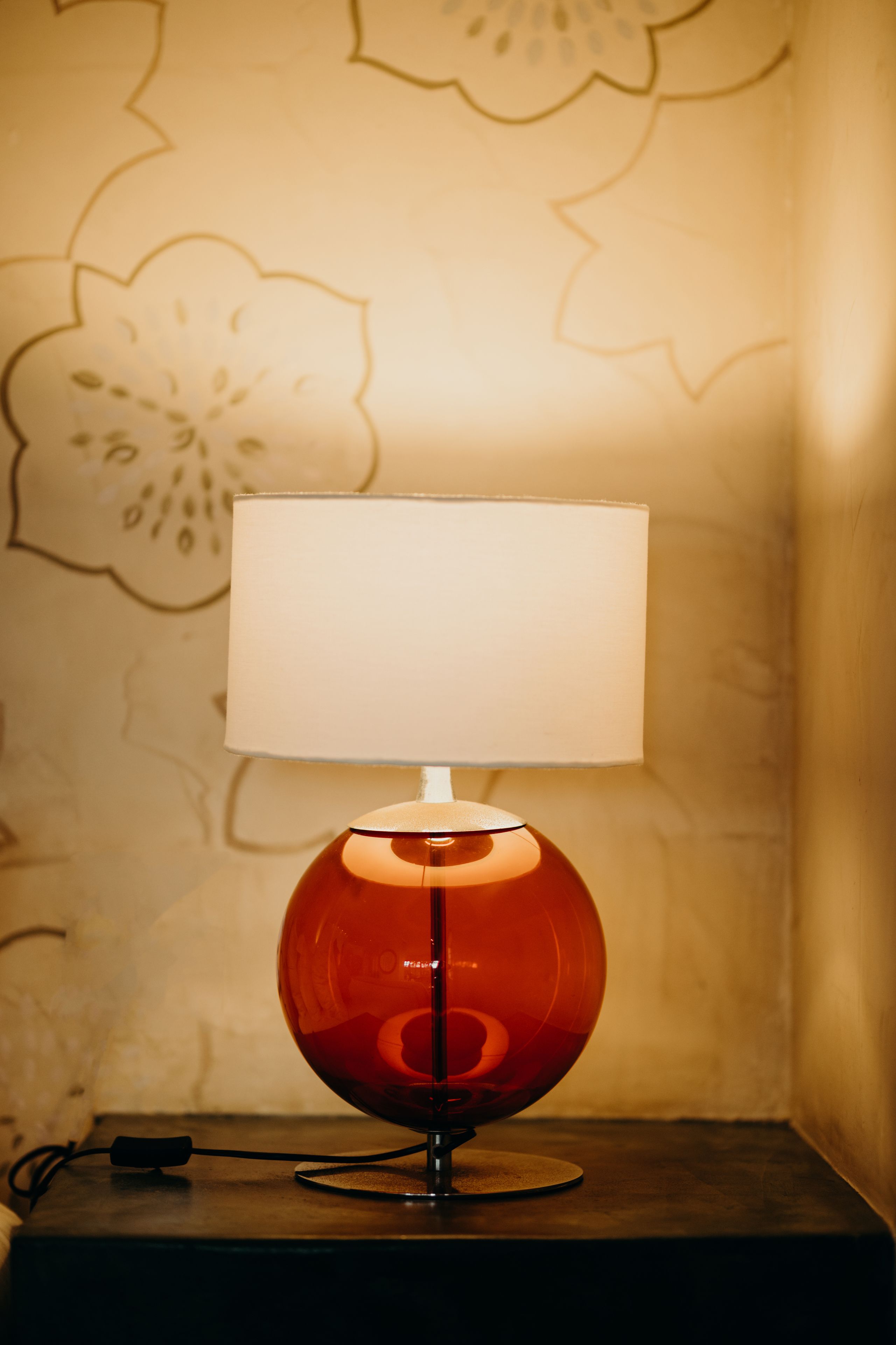 A table lamp with a white shade on a wooden table against a wall with floral wallpaper.