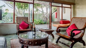 Una habitación con mesa de centro de vidrio, sillas de mimbre, un sofá con cojines rojos y grandes ventanales con vistas al exterior.