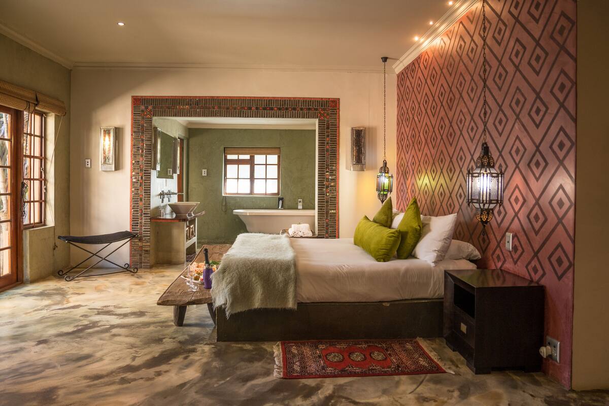 Een slaapkamer met een groot bed, een tafeltje, een bankje, een rood vloerkleed en uitzicht op de badkamer door een open deur.