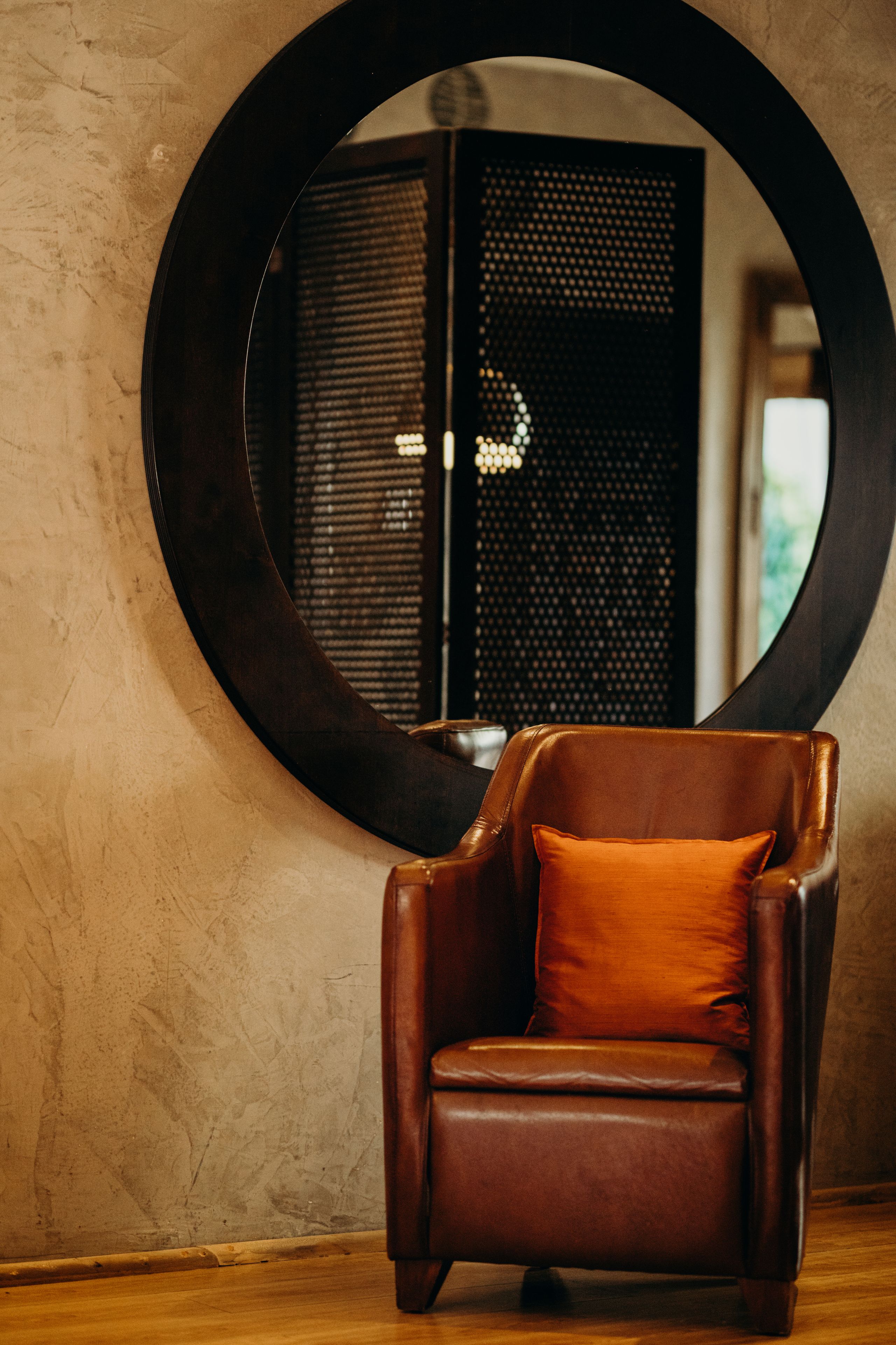 A leather armchair with an orange cushion in front of a round mirror on a textured wall.