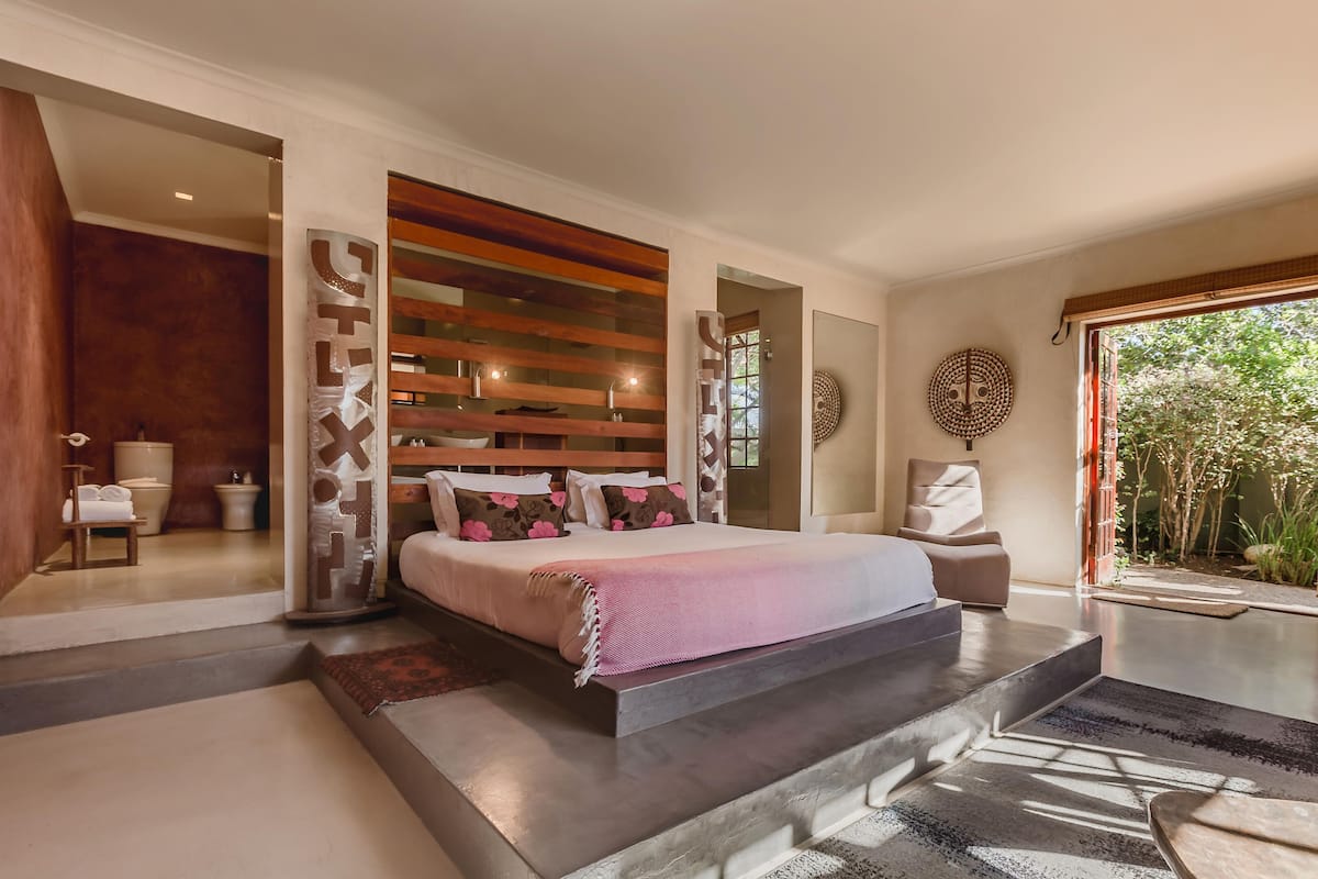 Een moderne slaapkamer met een groot bed, een houten lattenkap, een zithoek met een stoel en uitzicht op groen door een groot raam.