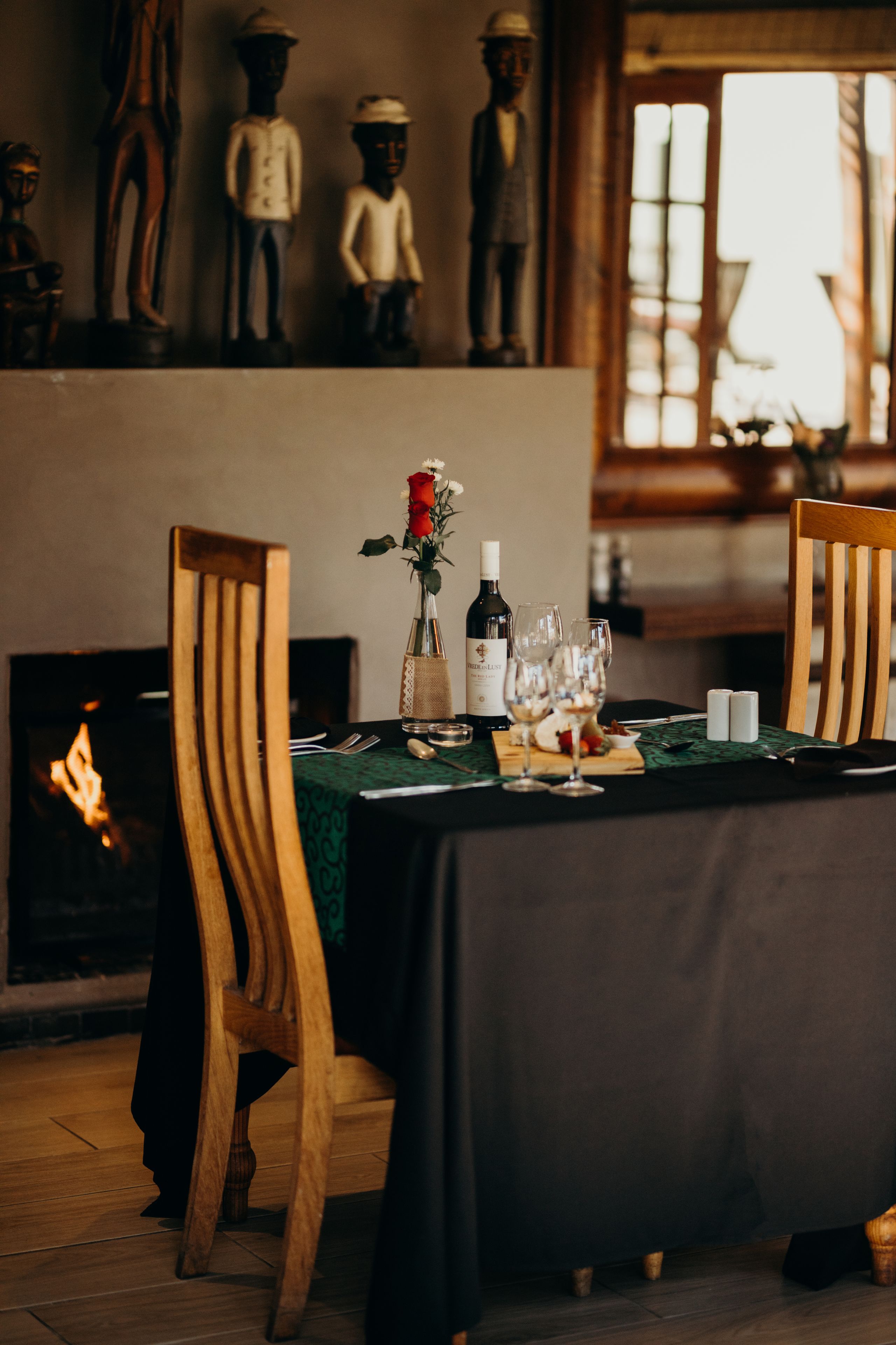 Una mesa de comedor dispuesta para dos personas, con mantel verde, sillas de madera, un florero con una rosa roja, copas de vino y una botella de vino.
