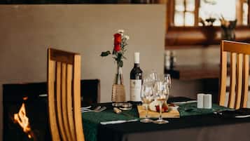 Una mesa de comedor dispuesta para dos personas, con mantel verde, sillas de madera, un florero con una rosa roja, copas de vino y una botella de vino.