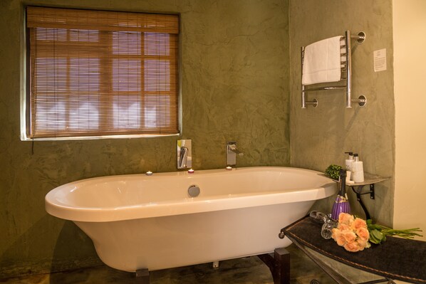 A bathroom with a freestanding bathtub, a window with blinds, a towel rack, and a small shelf with toiletries.