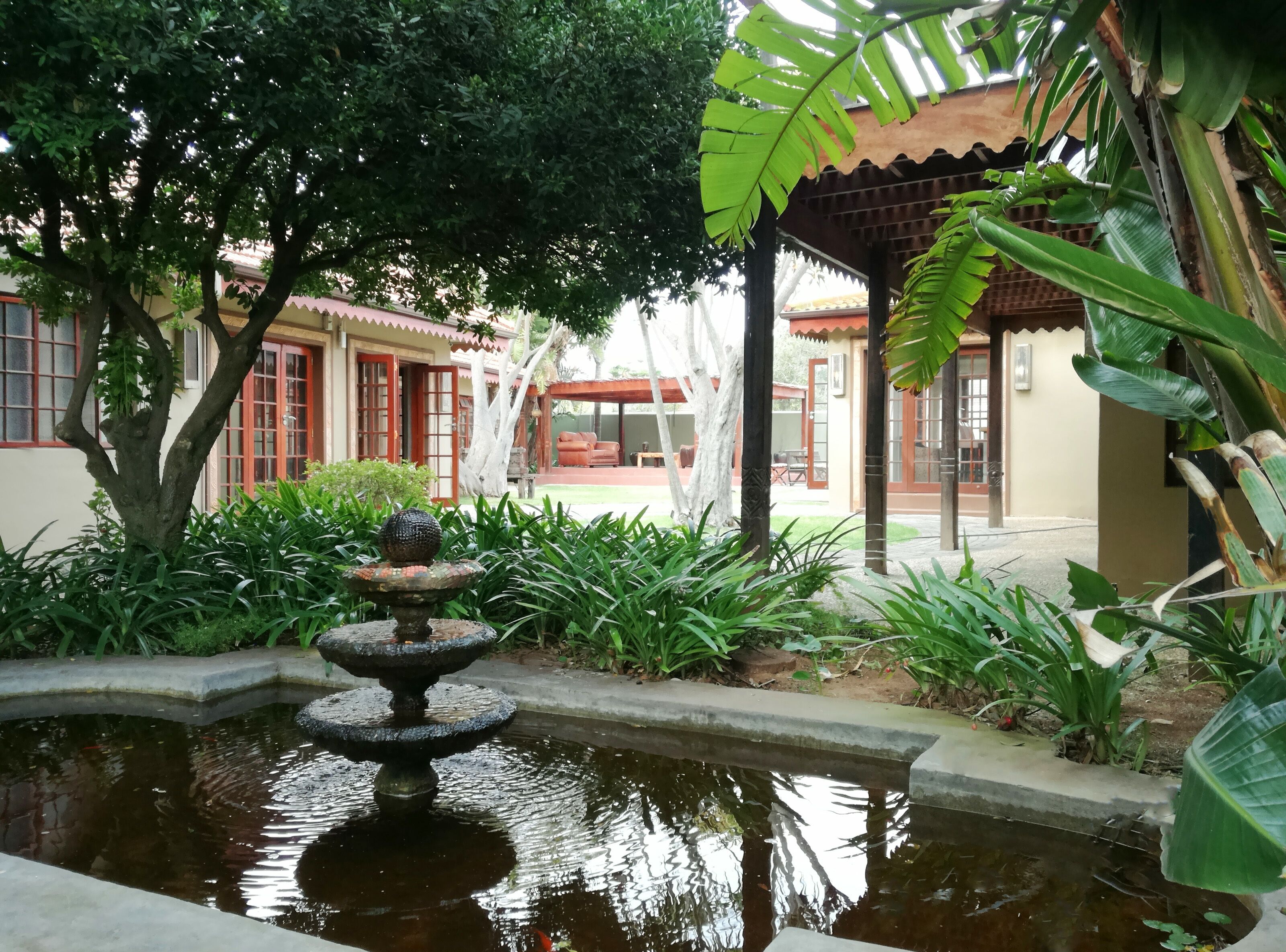 Un patio tranquilo con una fuente, abundante vegetación y un edificio tradicional con puertas rojas y techo de tejas.