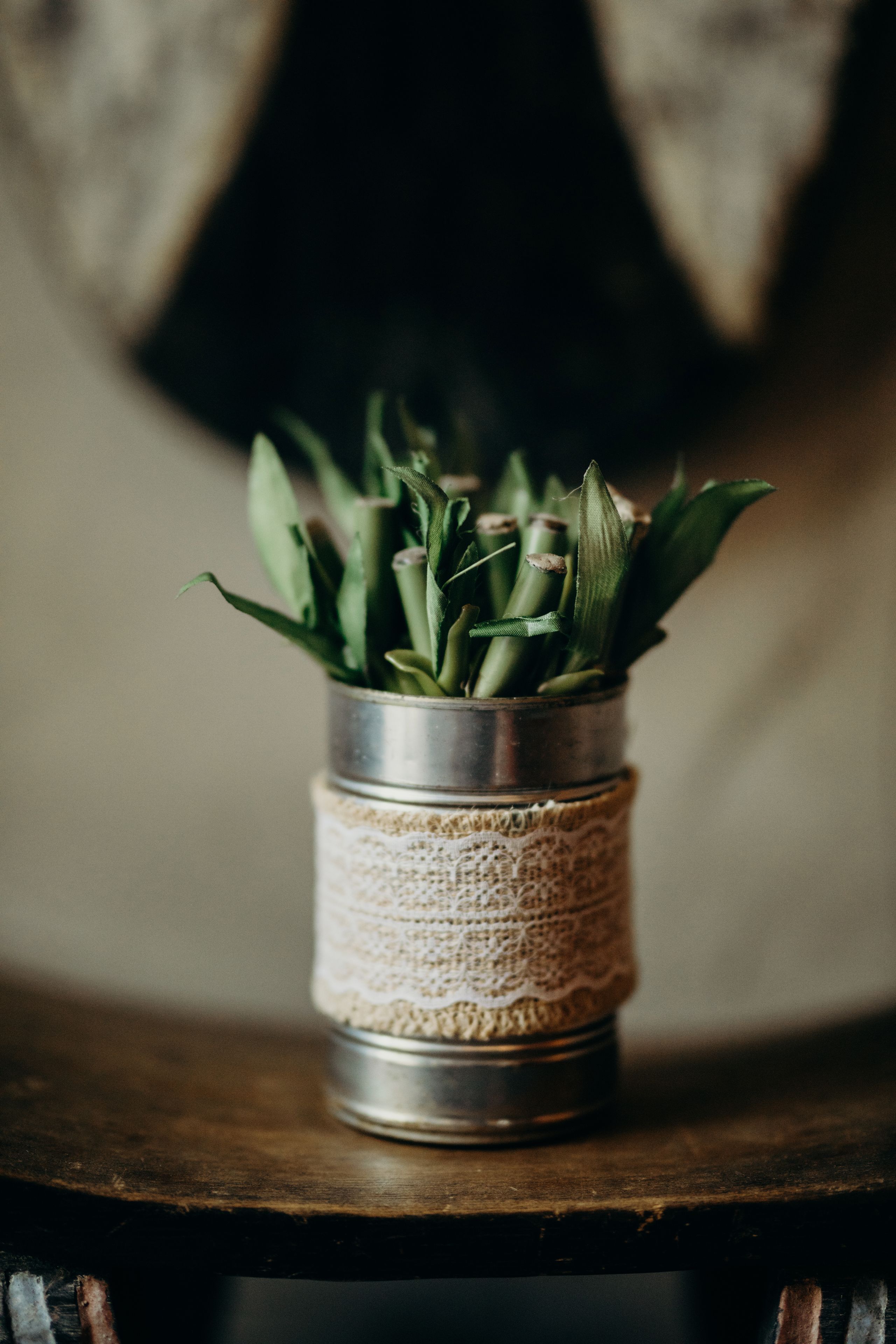 A metal container with a lace band, containing green plants, placed on a wooden surface.
