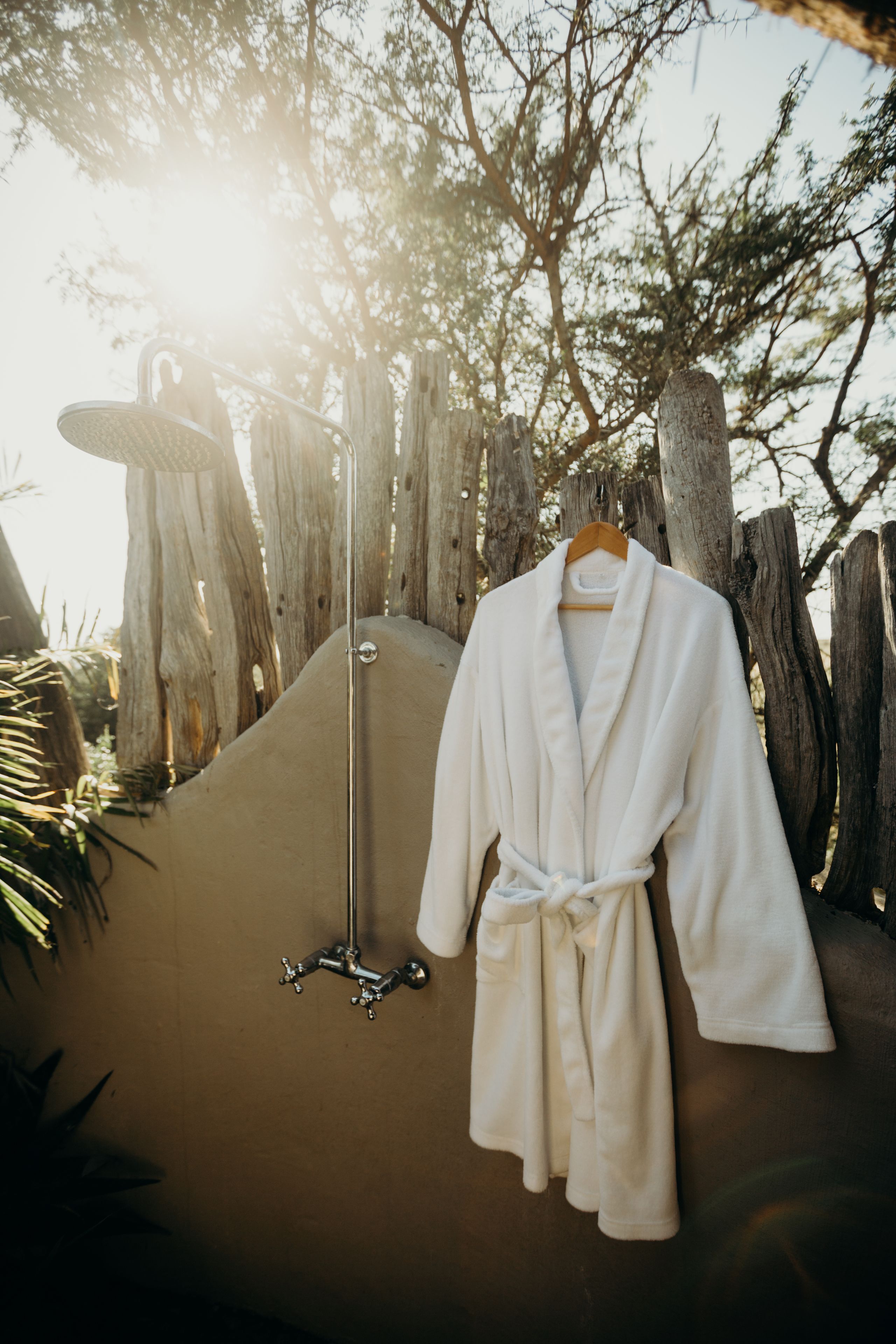 A white bathrobe hanging on a wooden hanger, a showerhead, and a wooden fence with trees in the background.