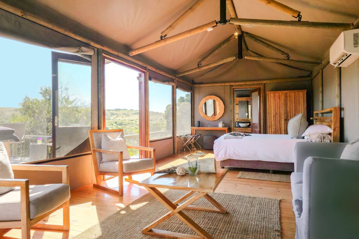 Una habitación amplia con una cama grande, un escritorio de madera, una silla y un sofá. Amplias ventanas ofrecen vistas al exterior.
