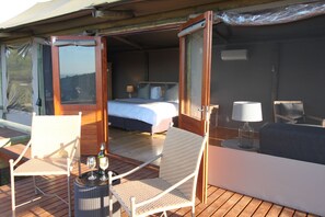 Une chambre ouverte avec un lit, deux chaises, une table avec une bouteille et un verre, et une vue sur l’extérieur.