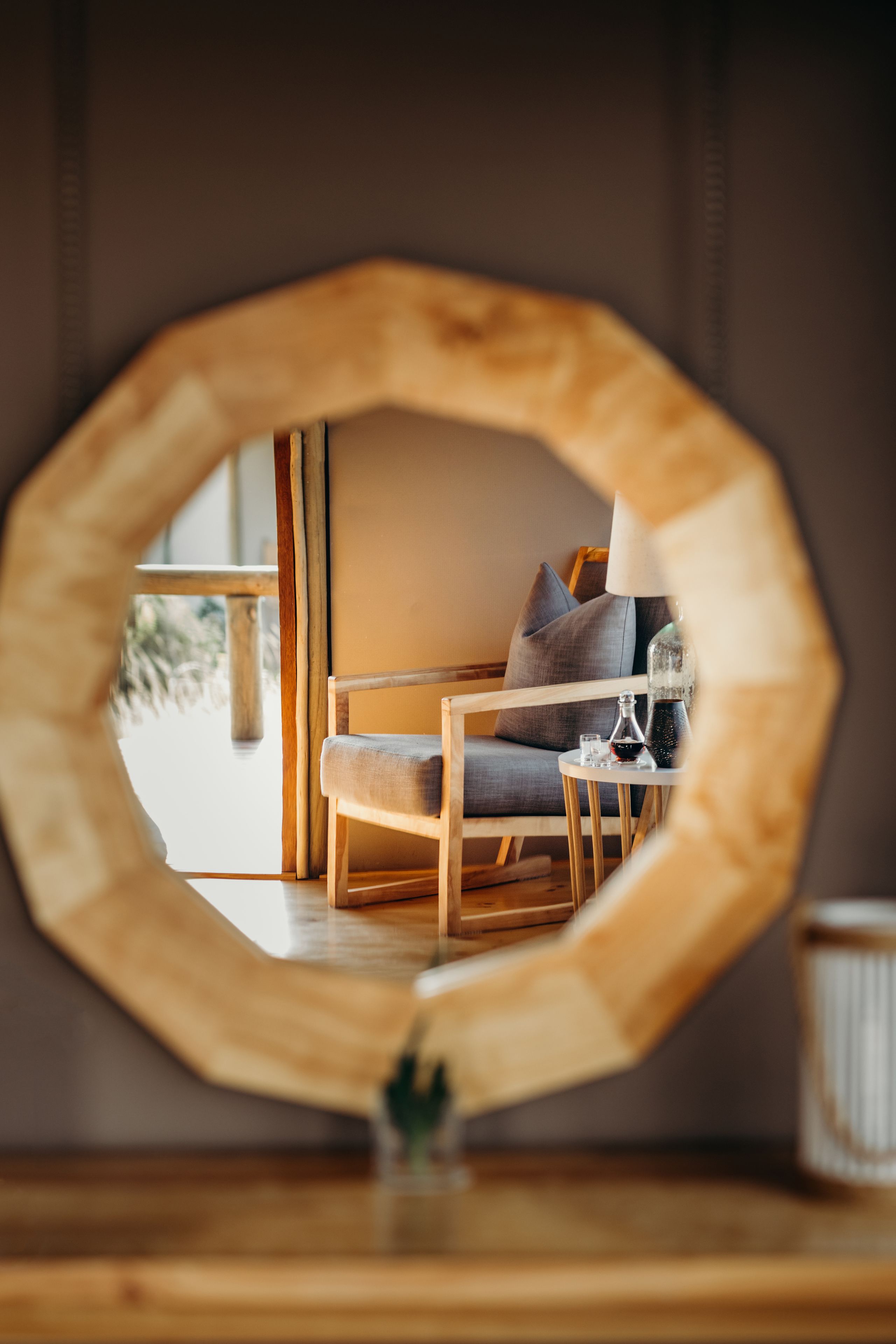 A round mirror reflecting a room with a wooden chair, a small table, and a lamp.