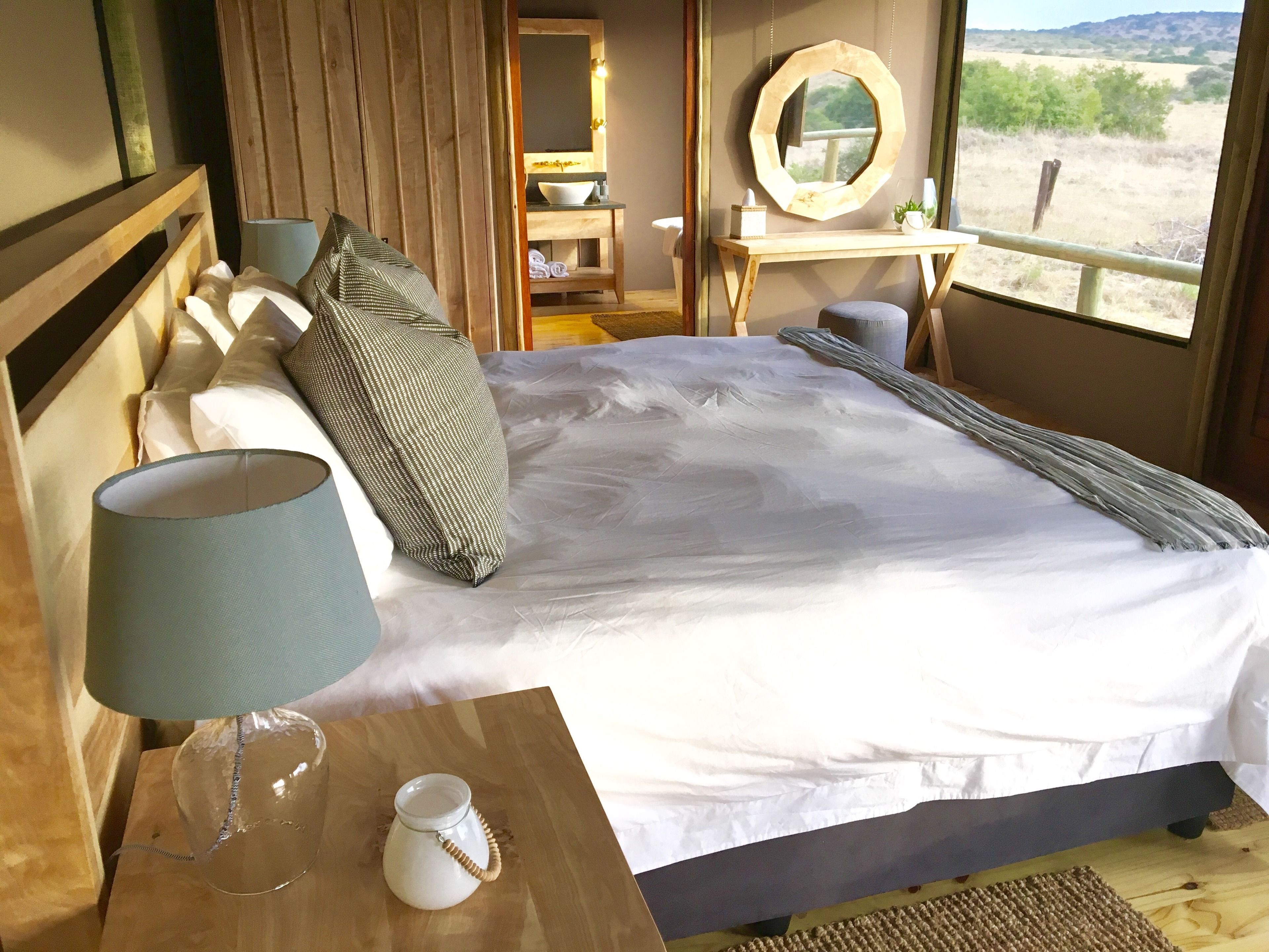 A neatly made bed with white linens, a bedside table with a lamp, and a view of a natural landscape through a window.
