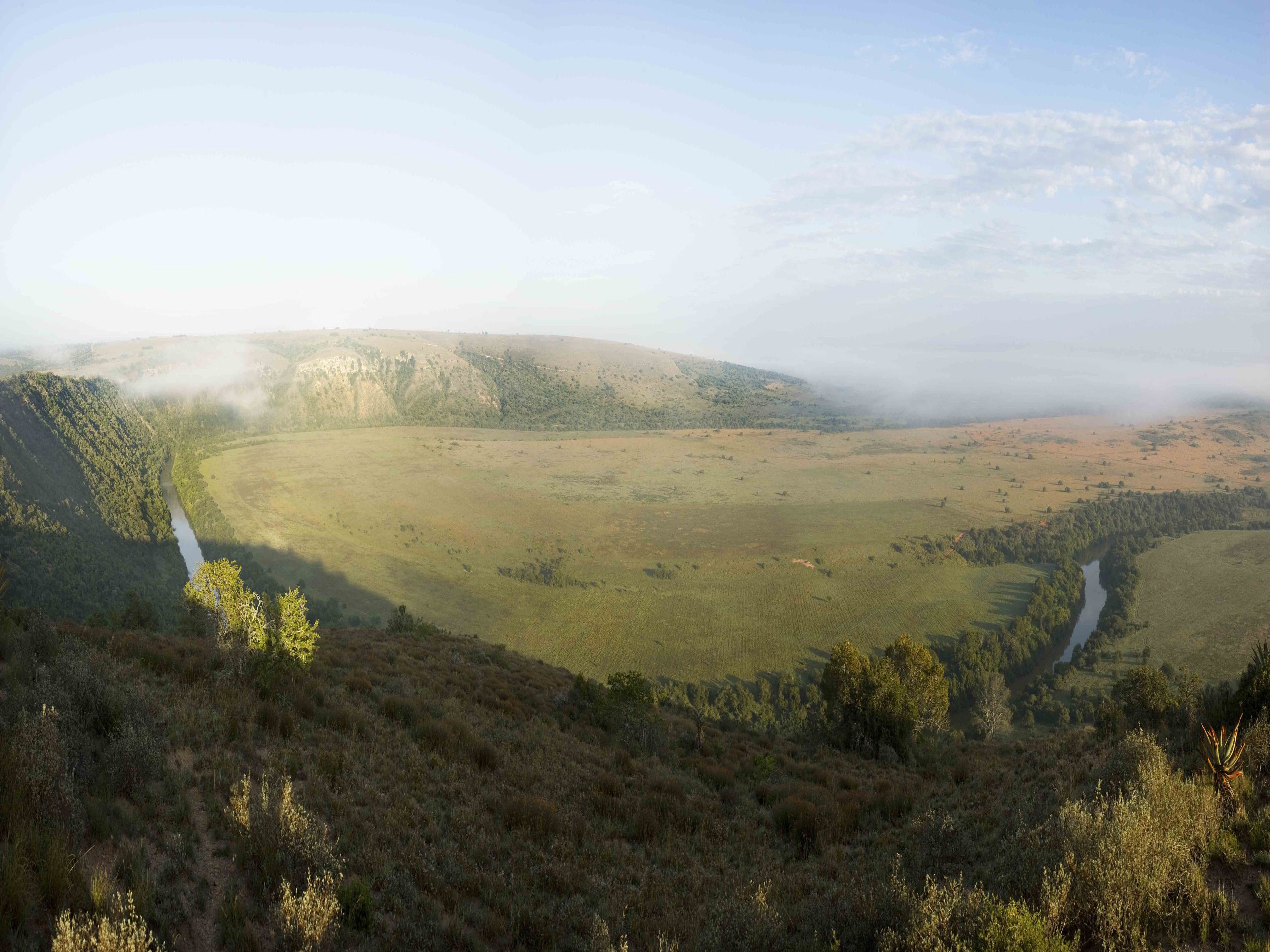 A panoramic view of a valley with a river, surrounded by hills and vegetation.