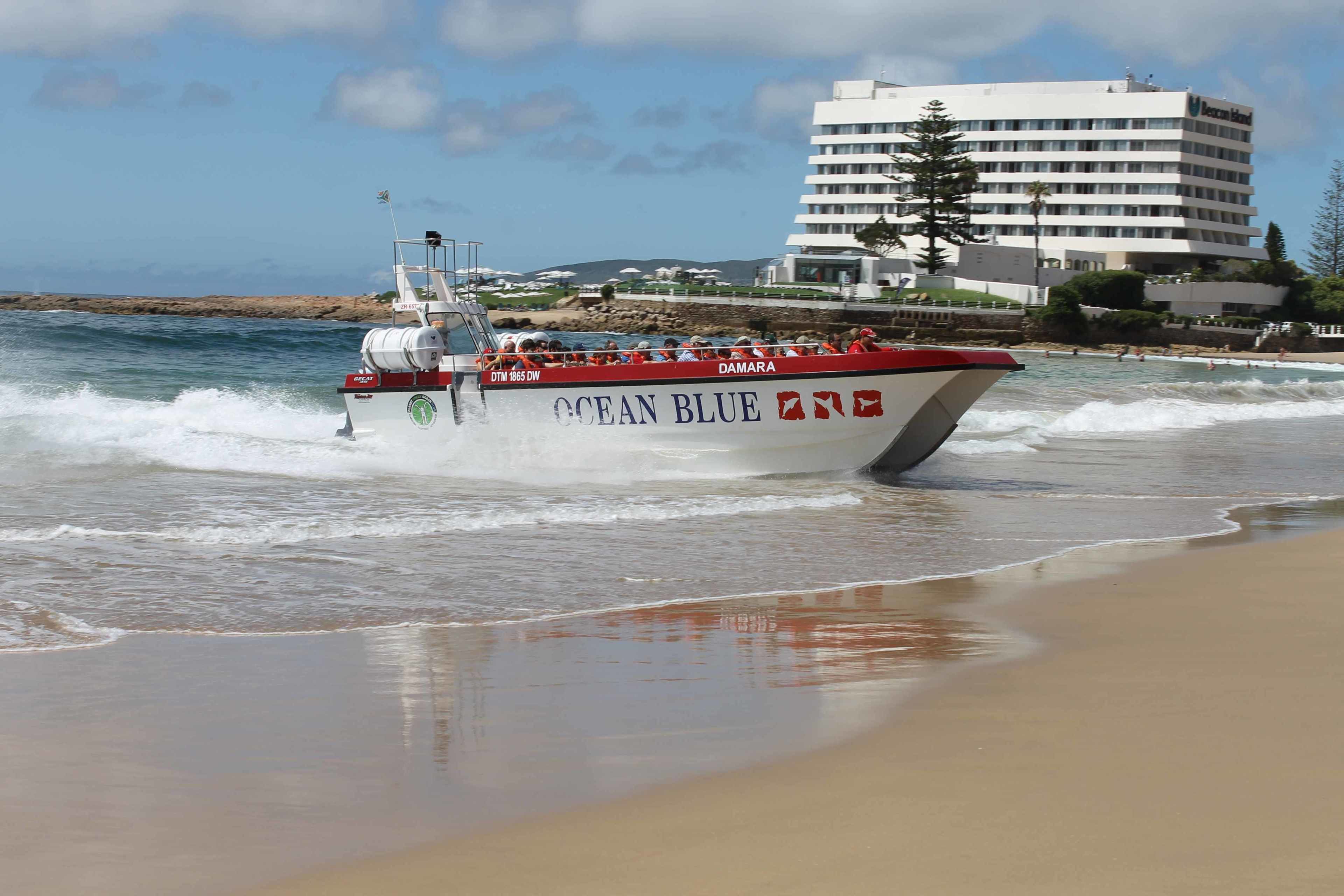 Un bote llamado 'Ocean Blue' con un diseño blanco y rojo, avanzando por una playa con olas. Al fondo, se observa un edificio blanco y de varios pisos, probablemente un hotel o resort.