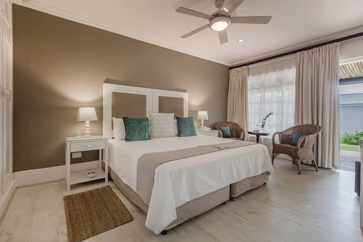 Une chambre avec un grand lit, deux tables de chevet avec des lampes, un ventilateur de plafond, un fauteuil en osier et une vue sur l’extérieur grâce à de grandes fenêtres.