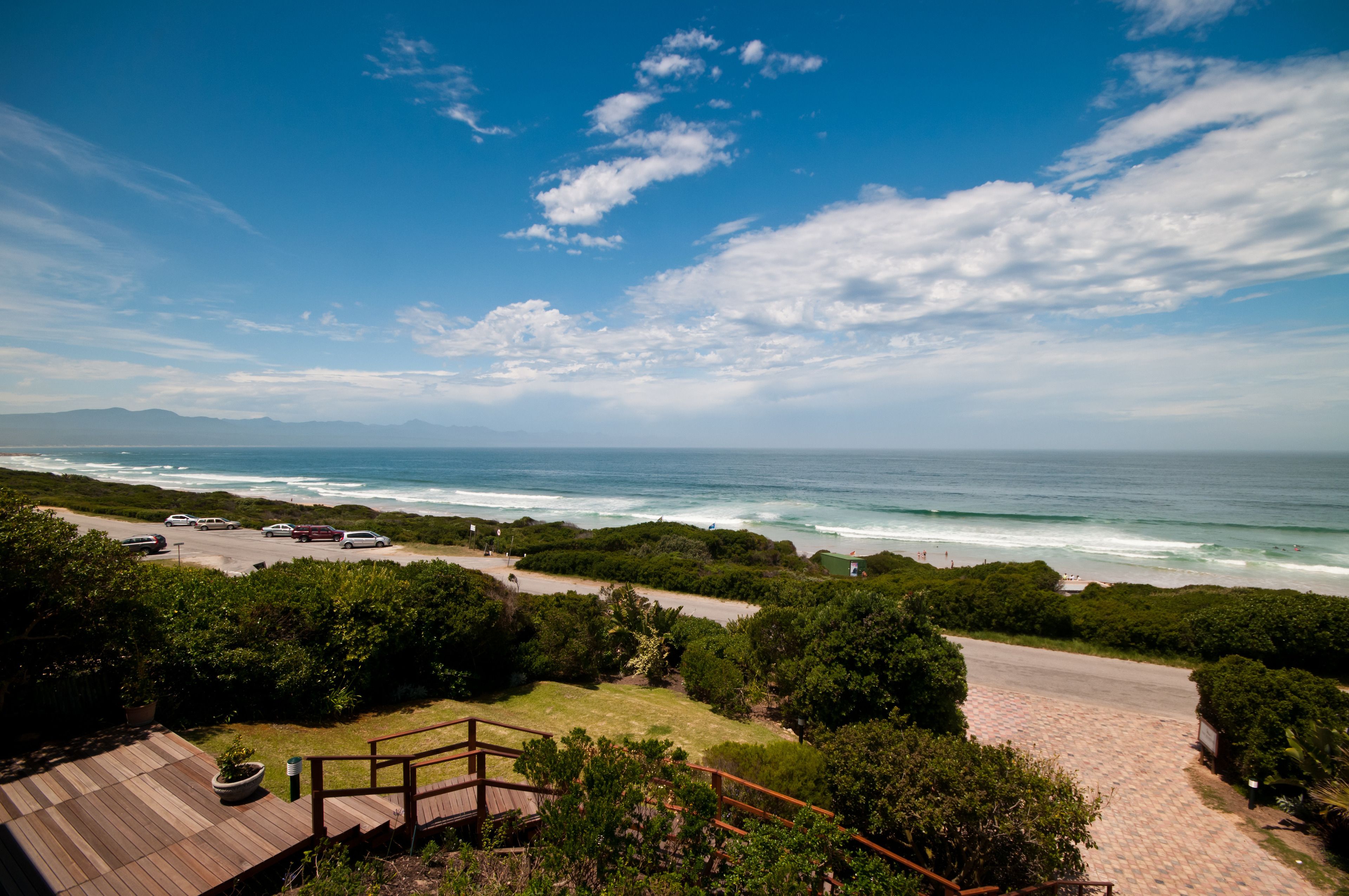 A coastal view with a wooden deck, a pathway leading to the beach, and a parking area with cars.
