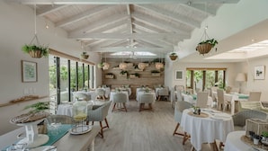 Une salle à manger spacieuse avec de hauts plafonds, des poutres en bois et des plantes suspendues. Les tables sont recouvertes de nappes blanches et entourées de chaises en osier.
